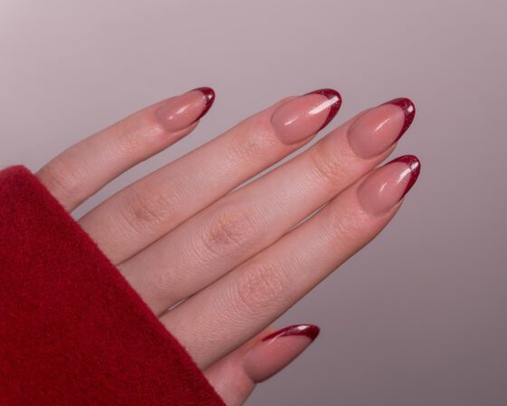 Diagonal close-up of an outstretched hand with red cuff on gray, showing medium blush almond nails with deep red French tips