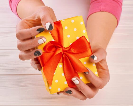 Two hands holding a yellow polka-dot gift with an orange bow on pale wood, showing short nails with charcoal, white and grey geometric colour blocks