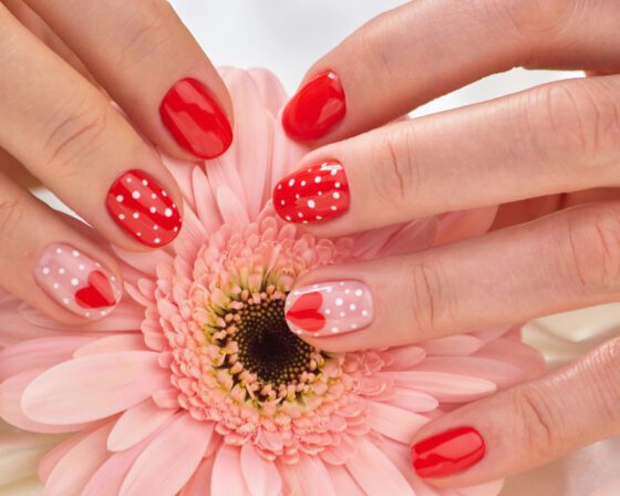 Close-up of several hands meeting around a pink flower, showing short bright red nails with white polka dots and pale pink accents with small red hearts