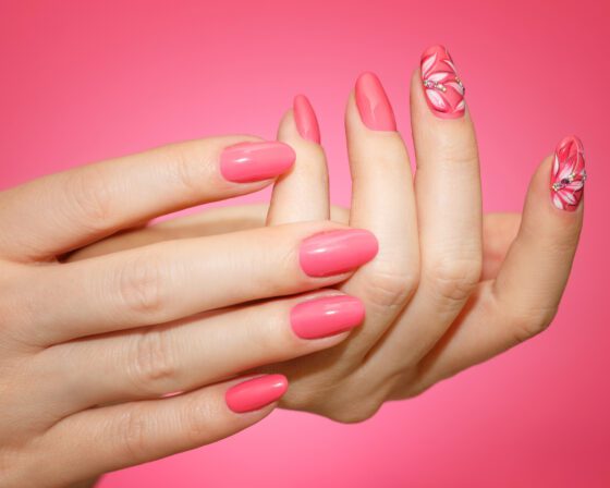 Posed hands against a saturated pink studio background, showing glossy coral pink almond nails with floral line art on accent nails