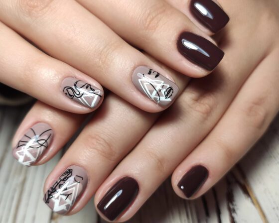 Close crop of crossed hands above pale wood, showing glossy deep plum nails with nude accents carrying black and white line art