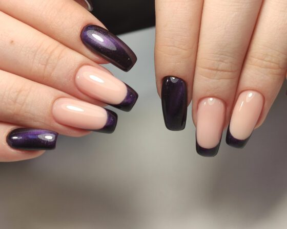 Close-up of two hands against pale gray, showing long nude nails with deep violet shimmer French tips