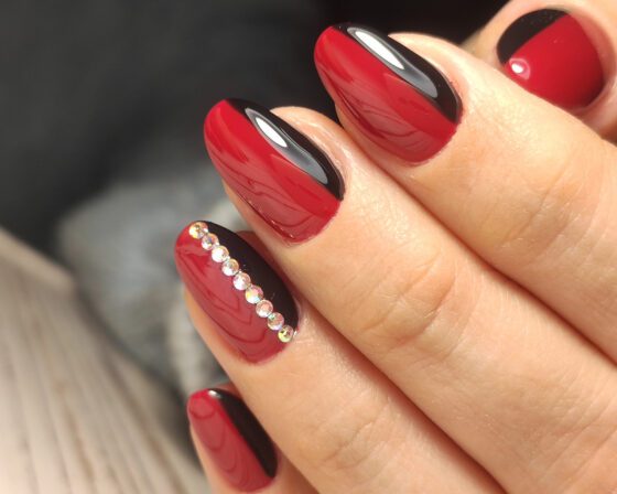Macro crop of fingers against a dark blurred background, showing glossy red nails with curved black side bands and a line of small crystals on an accent nail