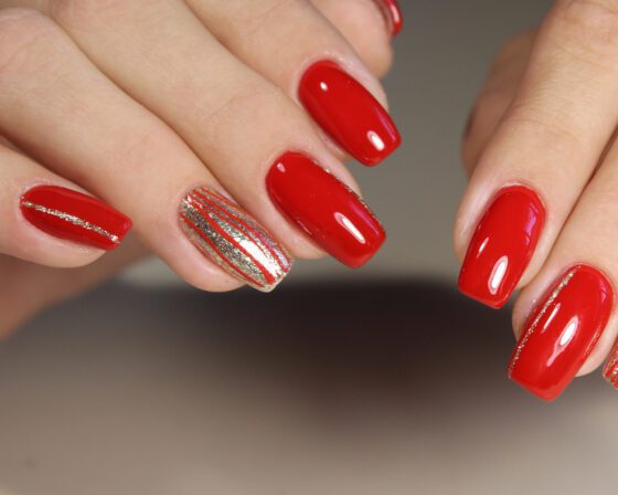 Two overlapping hands against a neutral gray backdrop, showing glossy bright red nails with silver-striped accent nails