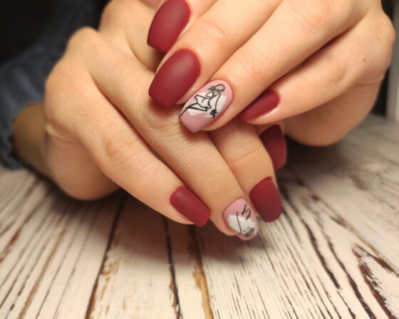 Clasped hands resting on pale wood, showing matte berry-red nails with a pale pink accent carrying a black line drawing
