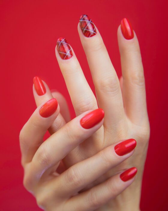 Posed hands against a saturated red studio background, showing medium glossy red almond nails with pale accents carrying fine silver and dark lines