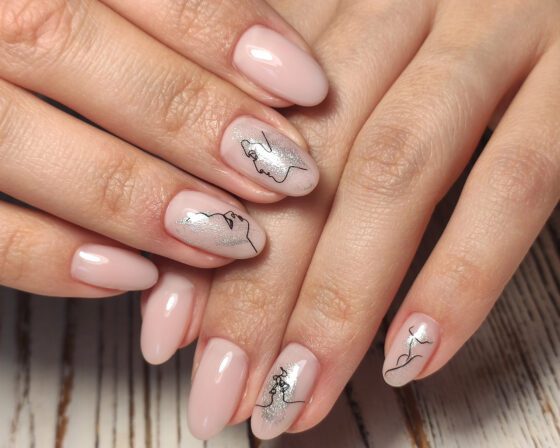 Extreme close-up of hands over pale wood, showing glossy pale pink almond nails with silver shimmer accents and fine black line drawings