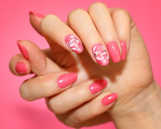 Centered close-up of one hand against a vivid pink studio background, showing glossy coral pink almond nails with floral line art on accent nails