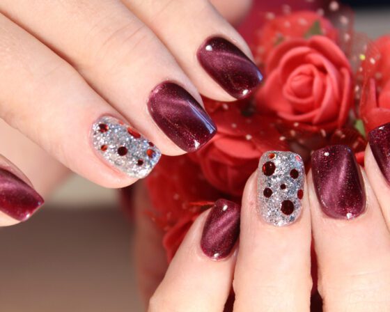 Close-up of hands in front of red flowers, showing wine red nails with a luminous sheen and a silver glitter accent set with small red and gold stones