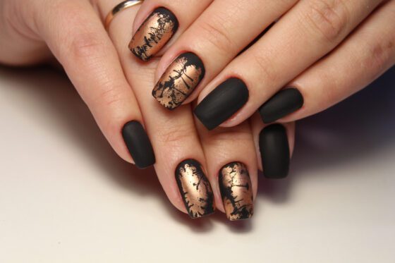 Close view of crossed hands on a pale neutral surface, showing matte black nails alternating with distressed metallic gold accents
