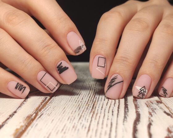 Close-up of several hands over a pale wood surface, showing short matte blush nails with black brush marks, outlined squares and fine white lines