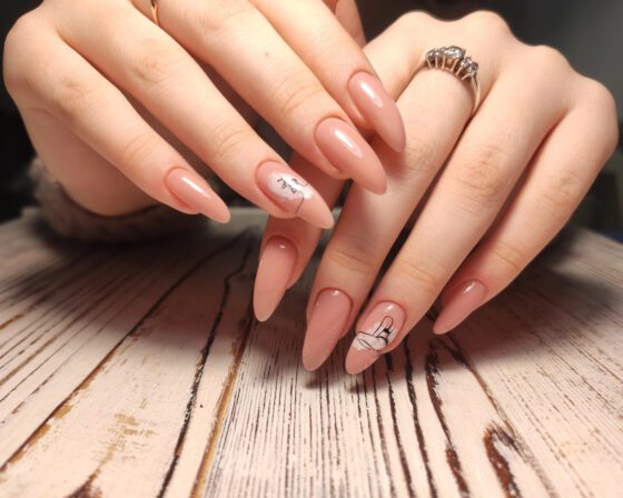 Close crop of overlapping hands above pale wood, showing long glossy peach-nude almond nails with small black line drawings on accent nails