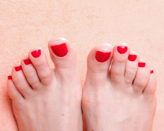 Wide frontal crop of both feet against a pink textured background, showing short toenails in a glossy bright red
