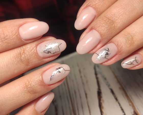 Close-up of crossed hands over pale wood, showing glossy pale pink almond nails with silver shimmer accents and fine black line drawings