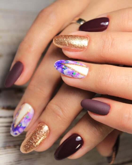 Portrait-oriented close-up of several hands against pale wood, showing almond nails mixing glossy and matte plum, copper glitter and violet-marbled white accents