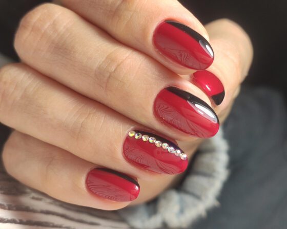 Close view of one hand with rings against pale wood, showing glossy red nails with curved black side bands and a line of small crystals on an accent nail