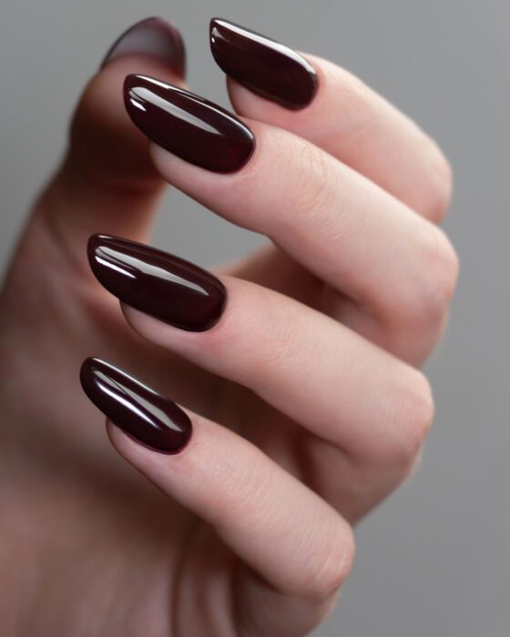 Vertical close-up of a curled hand against a gray background, showing medium almond nails in a glossy deep burgundy shade
