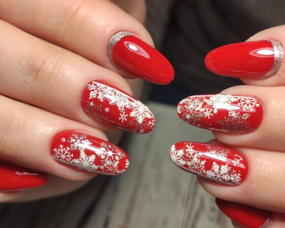 Close-up of overlapping hands on a pale textured surface, showing medium glossy red nails with white snowflake patterns on accents and silver sparkle near the base