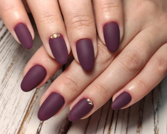 Close-up of crossed hands over pale wood, showing medium matte plum almond nails with small gold crystals near the base of accent nails