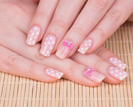 Close-up of overlapping hands above a woven mat, showing pale blush nails with white polka dots and small pink jewelled accents