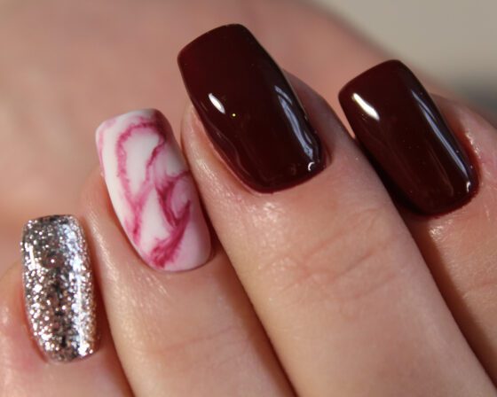 Extreme close-up of fingertips against pale skin-toned background, showing glossy burgundy nails with a pink-and-white marble accent and a silver glitter accent