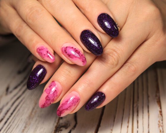 Tight horizontal close-up of two hands over pale wood, showing almond nails alternating bright pink marble and deep purple glitter