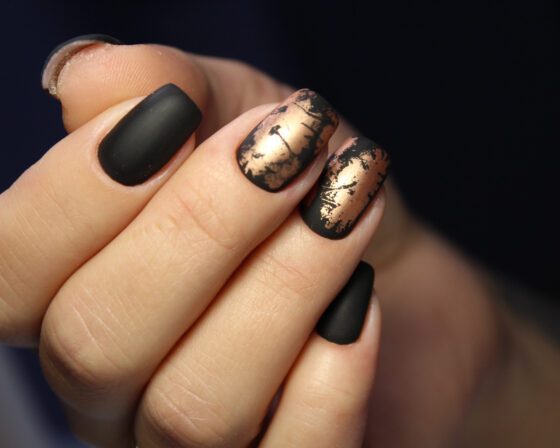 Extreme close-up of nail surfaces against a dark blurred background, showing matte black nails alternating with distressed metallic gold accents