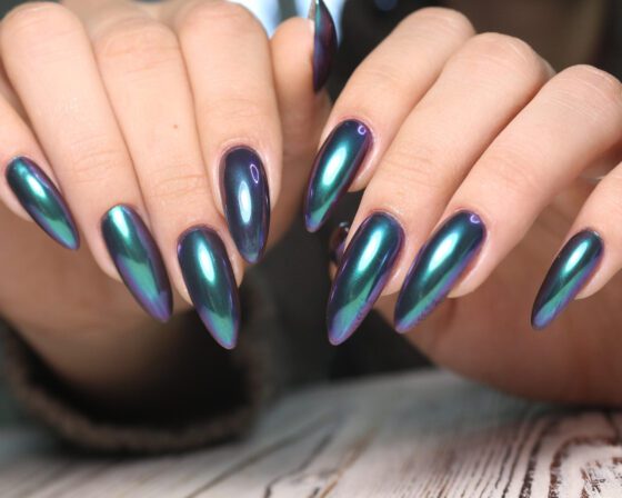 Close-up of two hands above a pale wood textured surface, showing long pointed navy nails with a turquoise and violet colour-shifting sheen