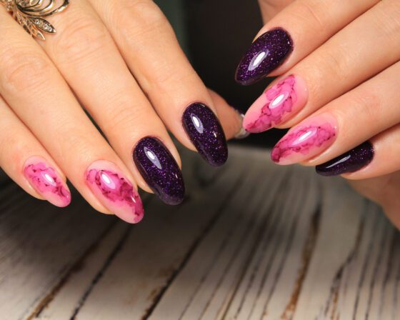 Close view of two hands above a pale wood surface, showing almond nails alternating bright pink marble and deep purple glitter