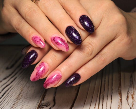 Close crop of hands against a pale wood surface, showing almond nails alternating bright pink marble and deep purple glitter