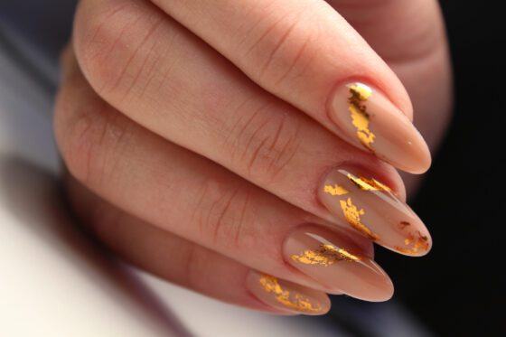 Tight close-up of fingers against a dark blurred background, showing medium sheer nude almond nails with irregular metallic gold flakes