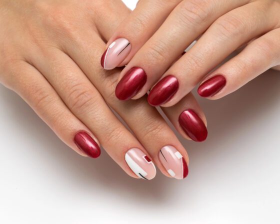 Close-up of overlapping hands on a bright neutral background, showing wine red metallic nails with nude accents carrying red and white geometric shapes