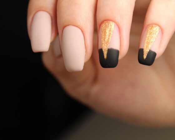 Macro view of raised fingertips against a dark blurred background, showing medium matte nude nails with black tips and gold glitter panels on accent nails