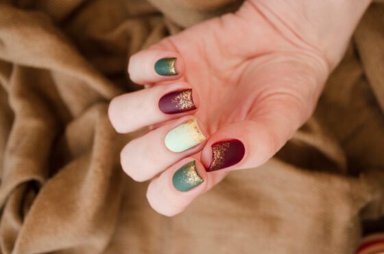 Close-up of curled hand in front of brown knit, showing short matte nails in forest green, wine and cream with gold glitter at the tips