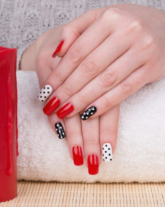 Vertical close-up of a hand against pale towel and woven textures, showing glossy red nails with black and white polka-dot accent nails