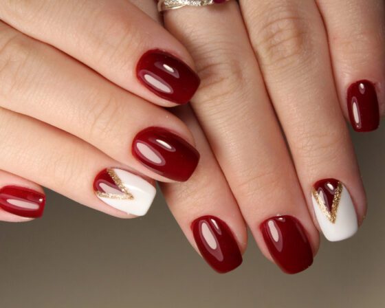 Macro close-up of fingertips against a neutral gray background, showing glossy burgundy nails with a white accent nail outlined by a fine gold V