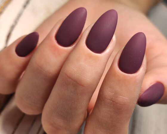 Macro crop of fingers against a softly patterned pale surface, showing almond nails in a muted matte plum