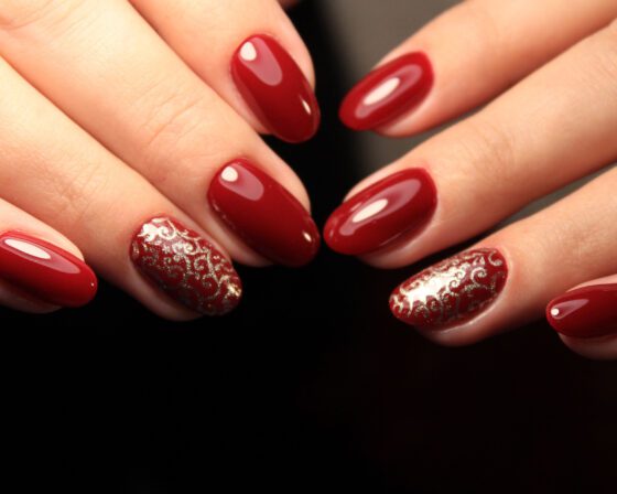 Tight view of several hands converging over a dark background, showing glossy deep red nails with gold filigree on one accent nail per hand
