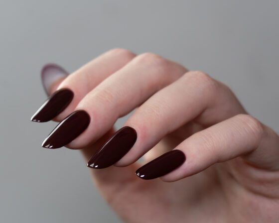 Close crop of several posed hands against a dark background, showing medium almond nails in a glossy deep burgundy shade