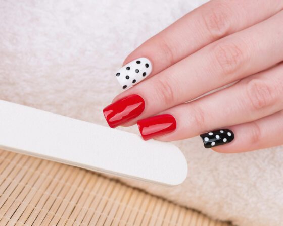 Close-up of a hand beside a white nail file on a soft towel, showing glossy red nails with black and white polka-dot accent nails