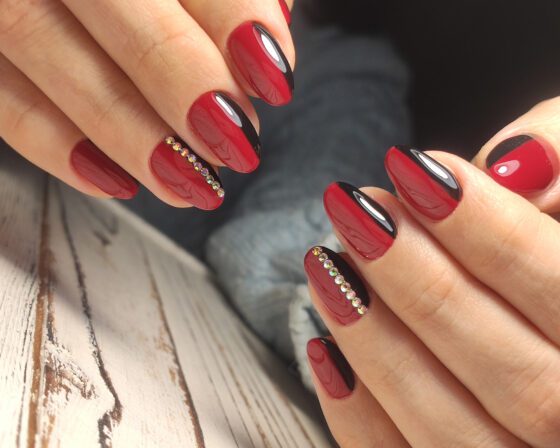 Close shot of two hands posed above a pale wood texture, showing glossy red nails with curved black side bands and a line of small crystals on an accent nail