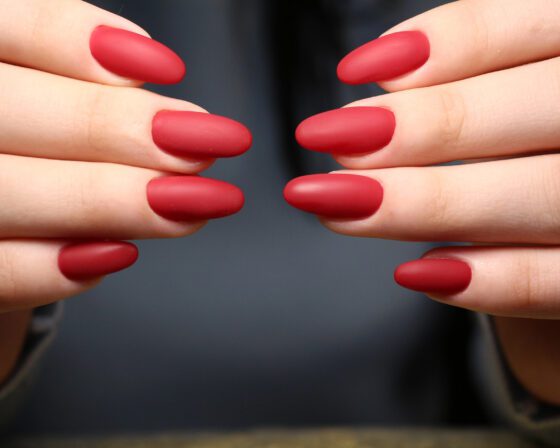 Two hands held toward each other, showing medium-length matte red nails against a dark background