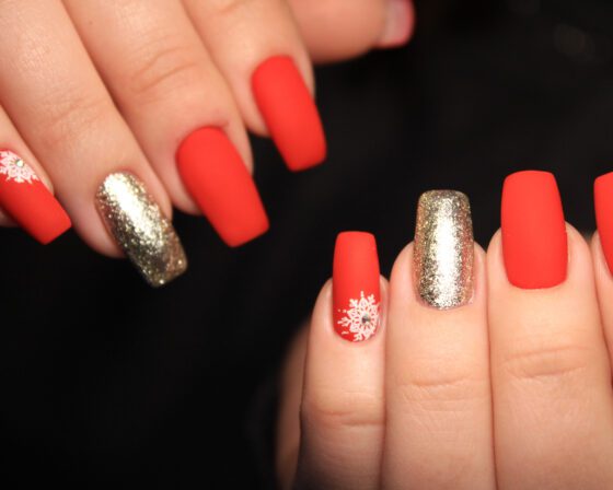 Tight close-up of hands against a dark background, showing long matte coral-red nails with a champagne glitter accent and a small white snowflake