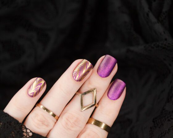 Close-up of hand with rings against black lace, showing short violet shimmer nails with gold chevron patterns on accent nails