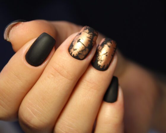 Extreme macro of fingertips against a dark blurred background, showing matte black nails alternating with distressed metallic gold accents