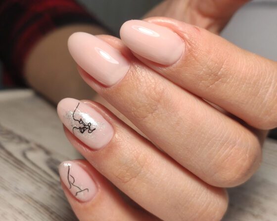 Tight close-up of hands with rings over pale wood, showing glossy pale pink almond nails with silver shimmer accents and fine black line drawings
