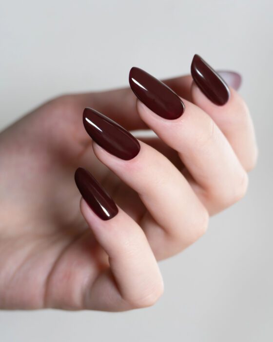 Vertical close-up of raised fingers against gray, showing medium almond nails in a glossy deep burgundy shade