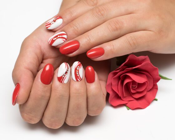 Hands held around a red rose against a bright background, showing glossy red nails and white accent nails with red swirls and small crystals