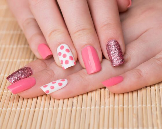 Wide close-up of two hands posed over a woven mat, showing hot pink nails with white polka-dot accents and rose-pink glitter accents