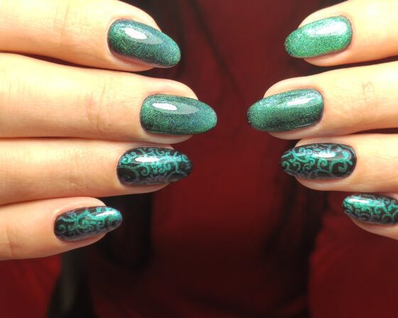 Symmetrical close-up of two hands facing each other against dark red, showing emerald green sparkle almond nails with dark accents carrying green scrollwork